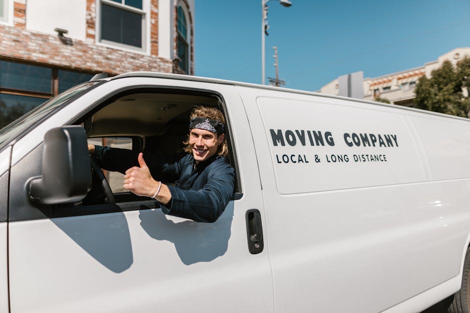 A young man with shoulder-length hair and a patterned bandana is sitting in the driver's seat of a white moving van, giving a thumbs-up gesture and smiling at the camera. The van belongs to Waterloo Man and Van, a company providing removals and furniture transport services, as indicated by the printed text on the side of the vehicle which reads 'MOVING COMPANY, LOCAL & LONG DISTANCE.' The van is parked outdoors on a street with modern buildings, blue sky, and utility poles visible in the background. The image captures the vehicle during part of the home relocation process, illustrating the loading or transporting phase of furniture and belongings, with the driver appearing confident in the move. The scene emphasizes aspects of packing, loading, and transporting household items efficiently, supported by the vehicle ready for a quick local move, in accordance with the tips for fast local moves as discussed in the webpage titled 'Lower Marsh market man and van tips for fast local moves, Waterloo' at waterloomanandvan.co.uk.