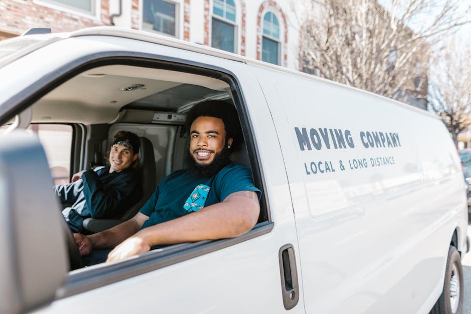 Inside a white moving van labeled with 'MOVING COMPANY' and 'LOCAL & LONG DISTANCE,' two individuals are seated. The man in the passenger seat, with dark curly hair and a beard, is smiling at the camera and wearing a blue shirt, suggesting he is part of the furniture transport or home relocation team from Waterloo Man and Van. The woman in the driver’s seat, with straight dark hair tied back and a headband, is also smiling, dressed in dark clothing. The van is parked outdoors, with a background of multi-storey buildings and leafless trees, indicating an urban environment. Visible in the interior are packing materials such as cardboard boxes, which are stacked and being prepared for loading or unloading, along with a few blankets or protective coverings likely used to safeguard furniture during transit. The scene depicts the loading process involved in a professional local or long-distance move, highlighting the logistics of packing, furniture transport, and vehicle loading typical in house removals.