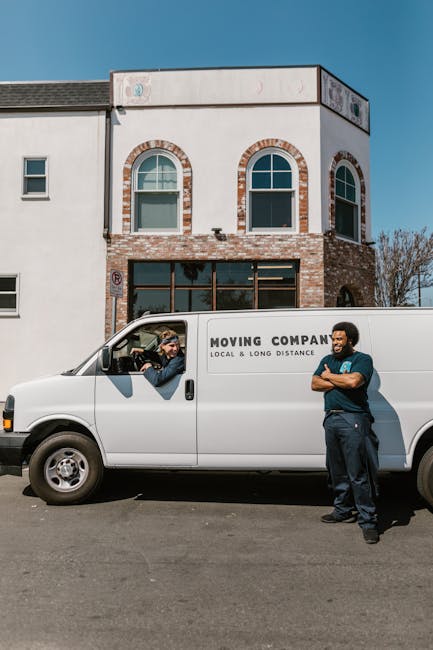 A young man with shoulder-length hair and a patterned bandana is sitting in the driver's seat of a white moving van, giving a thumbs-up gesture and smiling at the camera. The van belongs to Waterloo Man and Van, a company providing removals and furniture transport services, as indicated by the printed text on the side of the vehicle which reads 'MOVING COMPANY, LOCAL & LONG DISTANCE.' The van is parked outdoors on a street with modern buildings, blue sky, and utility poles visible in the background. The image captures the vehicle during part of the home relocation process, illustrating the loading or transporting phase of furniture and belongings, with the driver appearing confident in the move. The scene emphasizes aspects of packing, loading, and transporting household items efficiently, supported by the vehicle ready for a quick local move, in accordance with the tips for fast local moves as discussed in the webpage titled 'Lower Marsh market man and van tips for fast local moves, Waterloo' at waterloomanandvan.co.uk.