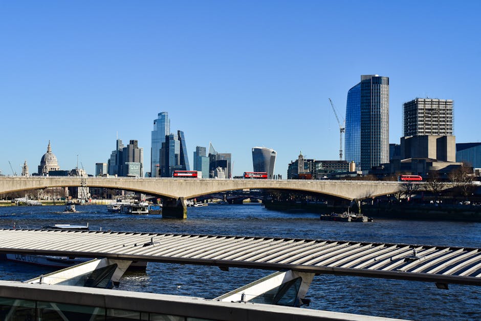 A clear daytime view of the London skyline across the River Thames, featuring the Millennium Bridge in the foreground with its sleek metal design and glass canopy, partially visible at the bottom of t