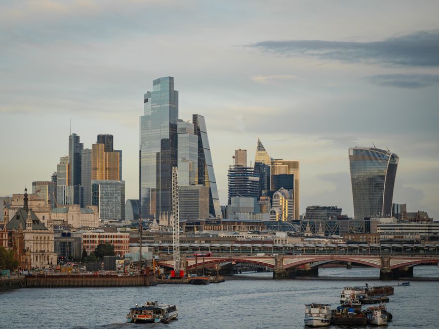 A clear daytime view of the London skyline across the River Thames, featuring the Millennium Bridge in the foreground with its sleek metal design and glass canopy, partially visible at the bottom of t