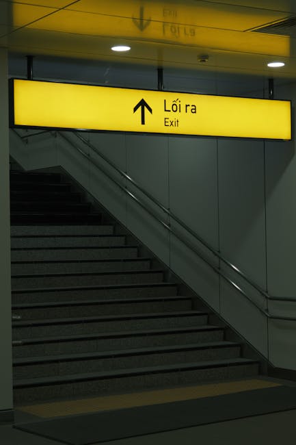 A staircase inside an indoor setting, likely within a transportation hub, leading upwards towards an exit. The staircase has dark-colored steps with a metal handrail on the right side. Above the stairs, there is a bright yellow illuminated sign with black text and an upward arrow indicating the direction to the exit, with the word 'Lối ra' in Vietnamese and 'Exit' in English. The surrounding walls are plain and light-colored, with additional lighting fixtures embedded in the ceiling. The scene is well-lit, and the environment appears clean and orderly, suitable for assisting home relocation and furniture transport processes as part of a house removals or moving service. Waterloo Man and Van may use such signage and staircases when planning efficient relocation logistics, especially in areas with challenging stair moves typical of certain buildings or stations.