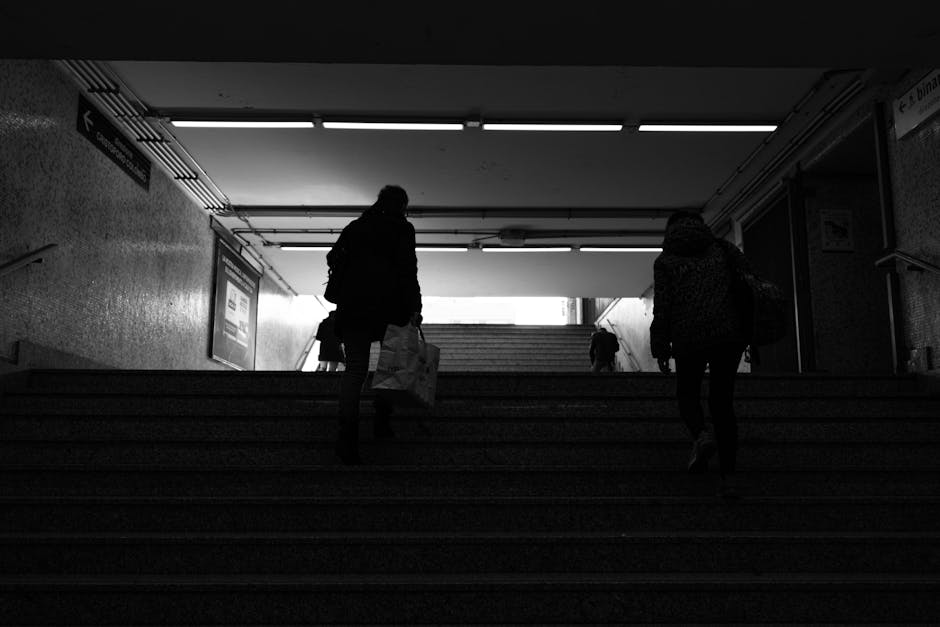 Inside a subway or train station, the image shows two people ascending a staircase leading to the surface, with a bright light or outdoor environment visible at the top. One person on the left is carrying a plastic shopping or moving bag and appears to be dressed in dark clothing, while the person on the right is wearing a coat with a patterned scarf, also carrying a bag. The staircase is divided into multiple steps made of textured material, with handrails on either side. The station interior features wall-mounted signage and overhead lighting fixtures, creating a contrast between the dimly lit interior and the natural light outside. Additional individuals can be seen at the upper landing, suggesting a common movement pattern typical during home relocations or journeys to new residences. This scene reflects common logistics involved in house removals, with the focus on passengers or movers transitioning between indoor and outdoor environments, often associated with packing, furniture transport, and the initial or final stages of a house move facilitated by professional services like Waterloo Man and Van.