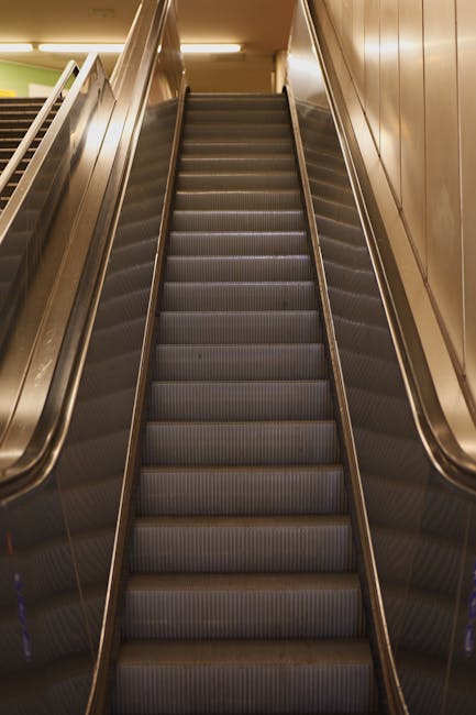 A staircase inside an indoor setting, likely within a transportation hub, leading upwards towards an exit. The staircase has dark-colored steps with a metal handrail on the right side. Above the stairs, there is a bright yellow illuminated sign with black text and an upward arrow indicating the direction to the exit, with the word 'Lối ra' in Vietnamese and 'Exit' in English. The surrounding walls are plain and light-colored, with additional lighting fixtures embedded in the ceiling. The scene is well-lit, and the environment appears clean and orderly, suitable for assisting home relocation and furniture transport processes as part of a house removals or moving service. Waterloo Man and Van may use such signage and staircases when planning efficient relocation logistics, especially in areas with challenging stair moves typical of certain buildings or stations.